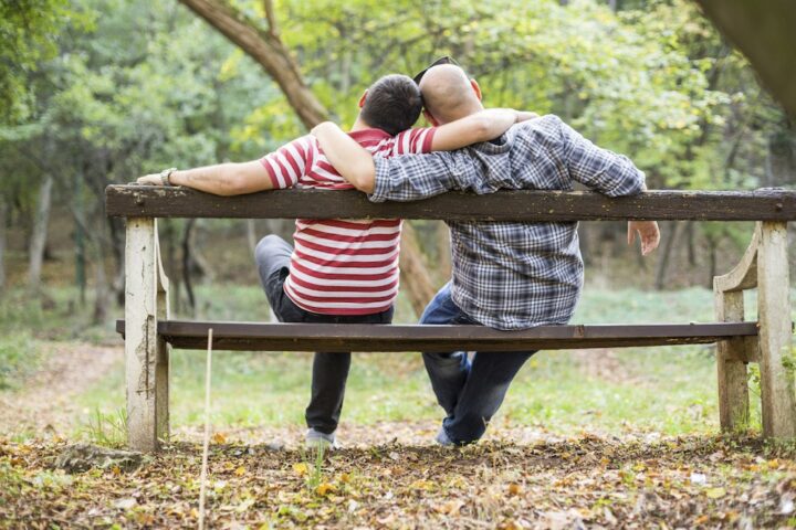 Two gay men sitting on a bench with their arms around each other looking at the forested park